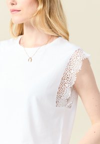 Valkoinen lyhythihainen paita, jossa on pitsireunus hartioilla. Pehmeää puuvillamateriaalia, jolla on sileä pinta ja pyöreä kaulus.