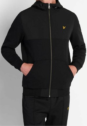 Person trägt einen schwarzen Zip-Up-Hoodie und passende schwarze Hose mit gelben Vogel-Logos, Hände in den Taschen des Hoodies, vor einfachem Hintergrund.