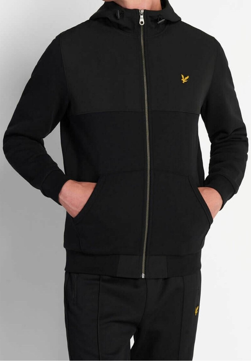 Personne portant un sweat à capuche noir zippé et un pantalon noir assorti avec des logos d'oiseaux jaunes, mains dans les poches du sweat, sur un fond uni.
