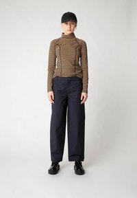 Top a maniche lunghe a righe in marrone e beige, abbinato a pantaloni larghi blu navy e stivali neri, completato da un cappellino nero.