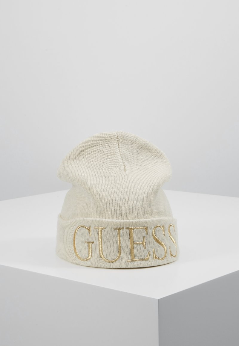 Bonnet tricoté couleur crème avec revers plié présentant un grand lettrage brodé en or "GUESS", exposé sur un socle blanc.