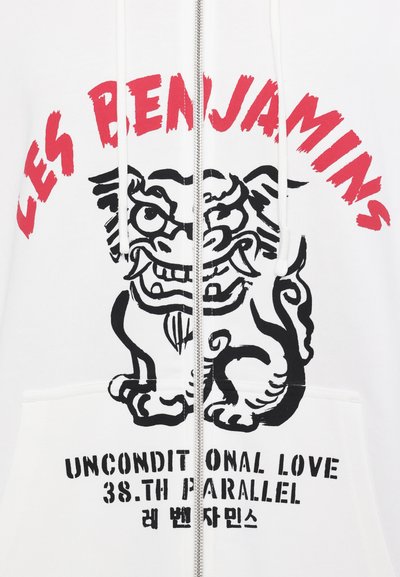 Λευκή ζακέτα με φερμουάρ που έχει μια μαύρη γραφική παράσταση μιας λιοντόμορφης φιγούρας και κόκκινο κείμενο που γράφει "LES BENJAMINS" και "UNCONDITIONAL LOVE 38.TH PARALLEL."