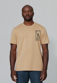 Beige katoenen t-shirt met een ronde hals en korte mouwen. Heeft een zwart grafisch ontwerp op de linkerborst.