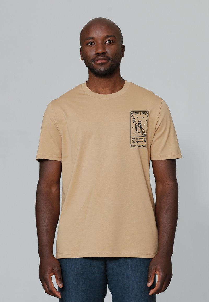Beige katoenen t-shirt met een ronde hals en korte mouwen. Heeft een zwart grafisch ontwerp op de linkerborst.