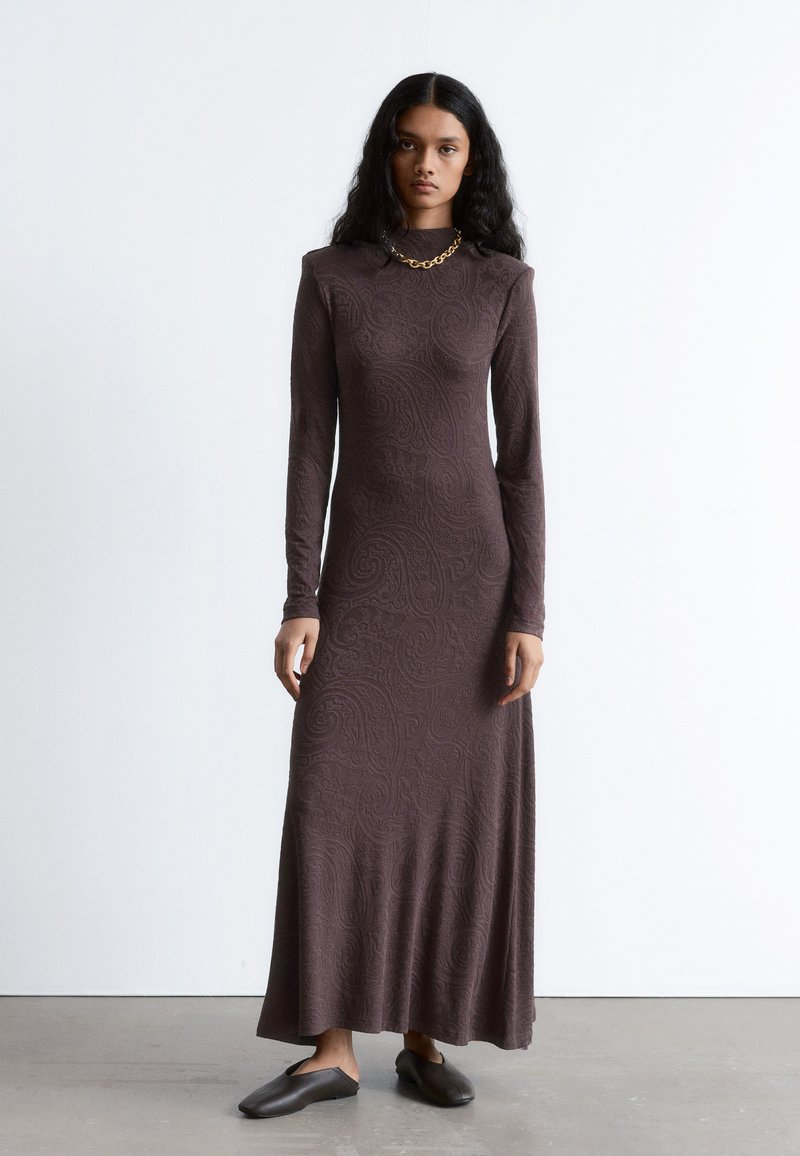Braunes, langärmliges Kleid mit einem bodenlangen Rock und Paisleymuster. Glatter Stoff, hoher Kragen und subtile Textur. Kombiniert mit dunklen Slippern.
