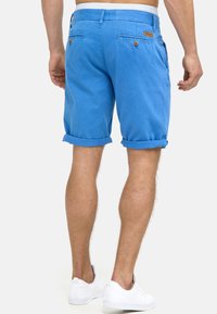 Homme portant un short bleu vif retroussé avec des poches arrière à boutons et des baskets blanches, vu de dos sur un fond uni.