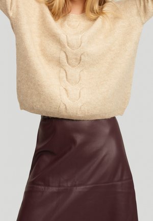 Beige kabelstrikket sweater med ballonærmer kombineret med mørkebrun læder nederdel i A-form på en stående person.