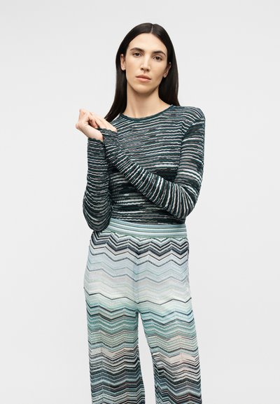 Žena s dlhými tmavými vlasmi, oblečená v pruhovanom top s dlhým rukávom a vysokopásových nohaviciach so vzorom cik-cak, stojaca pred obyčajným pozadím.
