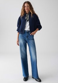 Giubbotto bomber navy con chiusura a zip, abbinato a jeans a vita alta di colore blu chiaro e stivali alla caviglia neri. Sciarpa a righe in accompagnamento.