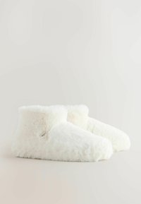 Pehmed, valged, karvased sääretasased sussid väikese riidest sildiga küljel, paigutatud lihtsa heleda tausta peale.