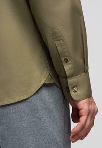 Poignet de manche d'une chemise kaki boutonée portée sur un pantalon gris avec une main visible détendue sur le côté.