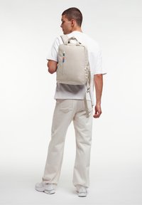 Zaino beige con un design strutturato, dotato di una tasca frontale con zip e un tirazip colorato. Indossato da una persona in una semplice camicia bianca.