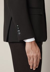 Ciemnobrązowy blazer o slim fit, z guzikami na rękawach i białą mankietą koszuli wystającą spod spodu.