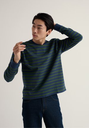 Jonge man die een blauwgroen gestreept shirt met lange mouwen verstelt, een donkere spijkerbroek draagt en voor een effen lichte achtergrond staat.