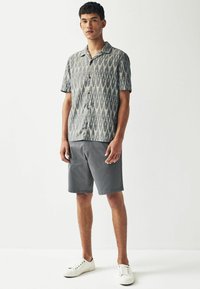 Kortærmet skjorte med knaplukning i et grå- og hvidt ikatmønster, parret med grå shorts. Modellen bærer hvide sneakers.