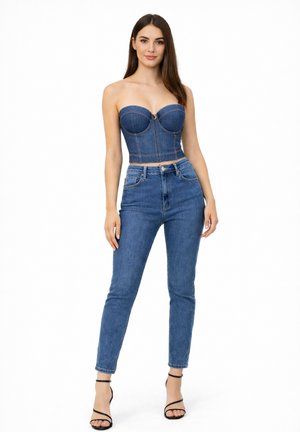Femme debout portant un corset en denim sans bretelles, un jean taille haute en denim et des talons noirs à brides sur fond blanc.