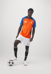 Camisa de futebol laranja e azul da Adidas, calções brancos, meias pretas até ao joelho com três riscas, de pé em cima de uma bola de futebol preta e branca.