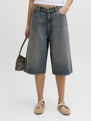 Person iført løstsittende, knelengde, falmede denimshorts, beige flate sko med stoffsløyfer, hvit kort topp, holder en liten mørk håndveske.