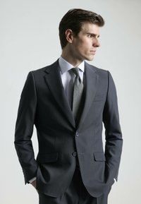 Traje gris oscuro con un corte entallado, diseño de un solo botón, solapas de muesca y una textura sutil. Combinado con una camisa de vestir clara y una corbata con estampado.