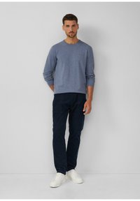 Mann, der an einer grauen Wand steht, wearing einen hellblauen Pullover, dunkelblaue Hosen und weiße Sneakers, mit neutralem Gesichtsausdruck nach vorne blickt.