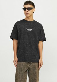 Sort t-shirt med korte ærmer og marmorering, med "Originals Studio" logo trykt på brystet. Sammen med lysebrune teksturerede bukser.