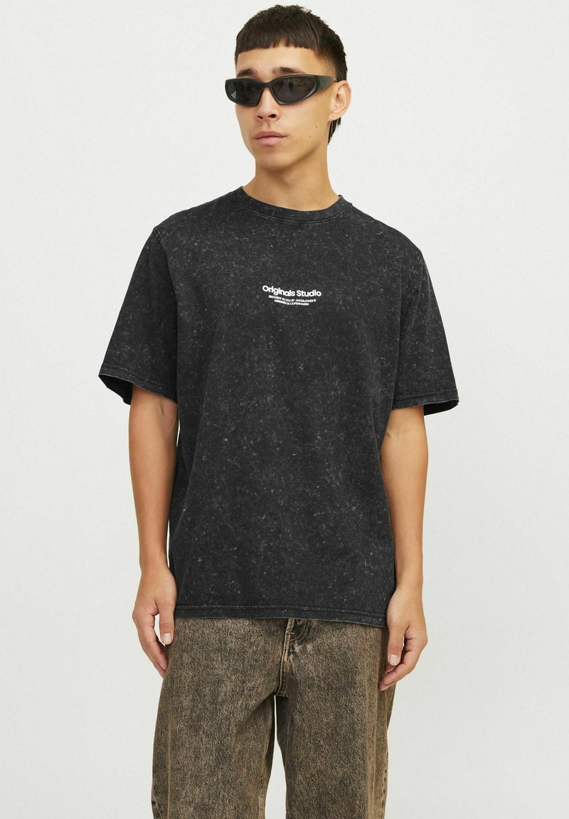 Sort t-shirt med korte ærmer og marmorering, med "Originals Studio" logo trykt på brystet. Sammen med lysebrune teksturerede bukser.