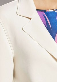 Witte gestructureerde blazer met een geknipt kraag, voorzien van een gladde stof, strakke lijnen en zichtbare stiksels. Een gebloemde blouse piept eronderuit.