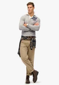 Camisa gris de manga larga con cuello blanco y logo de "Superdry State", pantalones beige y botas marrones con cordones; camisa a cuadros atada a la cintura.