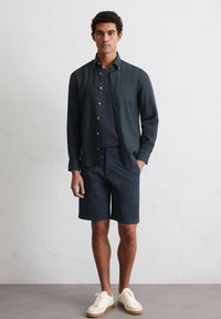 Donkerblauwe linnen button-up shirt over een marineblauw T-shirt, gecombineerd met marineblauwe shorts en witte sneakers. Het shirt heeft een casual pasvorm en zichtbare knopen.
