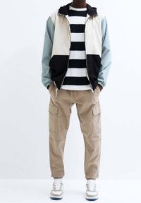 Mann in einer farblich abgesetzten Zip-Jacke, schwarzem und weißem gestreiften Shirt, beigen Cargo-Shorts und weißen Sneakers, der mit den Händen in den Taschen steht.