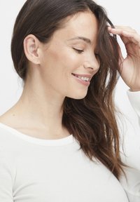 Weißes, geripptes Langarmoberteil mit weit ausgeschnittenem Kragen. Das Model hat lange, wellige braune Haare und berührt sanft ihre Haare.