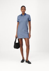 Robe polo bleue avec un motif géométrique, manches courtes et un col contrastant. Le mannequin tient un sac tissé noir et porte des mocassins noirs.