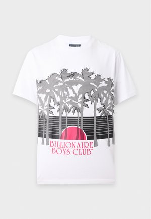 Jeune homme portant un t-shirt blanc à motif graphique avec des palmiers et un coucher de soleil, associé à un pantalon noir à jambes larges et des baskets noires et blanches.