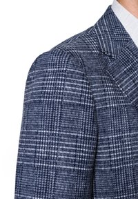 Zgarniany żakiet w granatowym i białym wzorze, z fakturowanym materiałem i szpiczastymi klapami. Zbliżenie na detal ramienia.