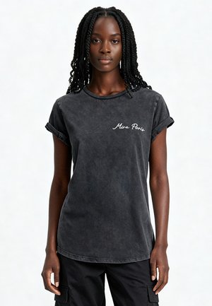 Jeune femme noire avec de longues tresses portant un t-shirt noir délavé avec « Mira Paris » écrit sur la poitrine, et un pantalon cargo noir.