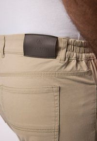 Pantaloni leggeri beige con elastico in vita e una patta in pelle marrone etichettata "BOSTON PARK"; presentano cuciture sottili e tasche.