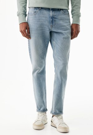 Lichtblauwe rechte jeans gedragen met witte sneakers en een lichtgroene longsleeve, te zien van taille tot voeten.