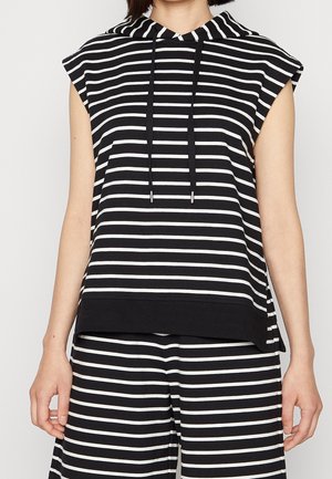 Černobílá bezrukávová mikina s horizontálními pruhy a šňůrkami, spárovaná s kalhotami se stejnými pruhy na osobě.