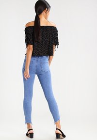 Schwarzes Off-Shoulder-Oberteil mit weißen Punkten, kombiniert mit hellblauen Skinny-Jeans mit fransigem Saum. Schwarze Blockabsatzsandalen.