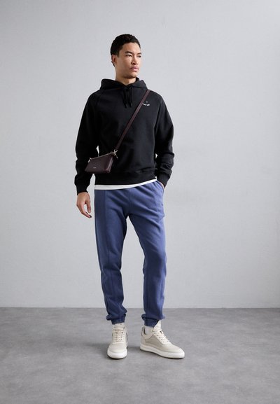 Zwarte hoodie met trekkoord, blauwe joggers en witte sneakers. Een kleine bruine crossbody tas wordt over de schouder gedragen. Eenvoudig ontwerp.