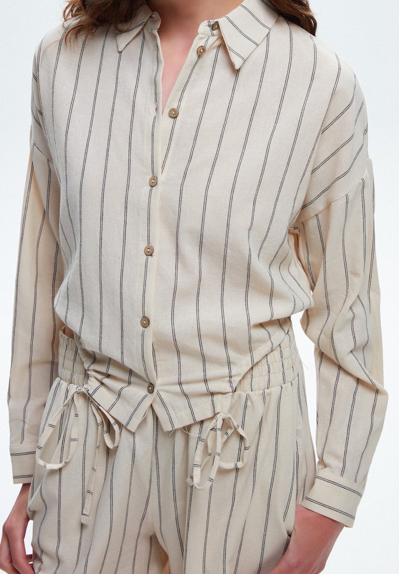 Femme portant une chemise beige à manches longues boutonnée et un pantalon assorti à cordon de serrage, avec de fines rayures verticales noires sur fond blanc.