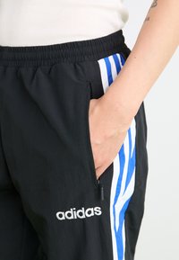 Sorte træningsbukser med elastisk talje, med lynlåslommer i siden, hvidt Adidas-logo og blå og hvide lodrette striber.
