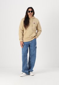 Beige hoodie with a front pocket and small logo patch, paired with light blue loose-fit denim jeans and white sneakers. Black sunglasses worn.