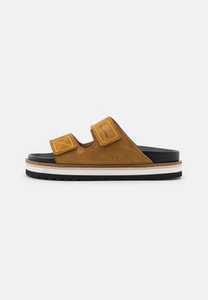 Brązowa zamszowa sandałka typu slip-on z dwoma żółtymi przeszyciami na rzepach i warstwową, grubą podeszwą w kolorze czarnym, białym i beżowym.