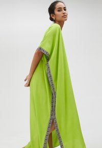 Grön kaftan med en flödande silhuett; har intrikata svart och vit kant längs de öppna sidorna och ärmarna för extra detalj.