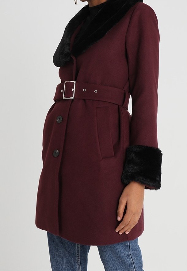 Abrigo burdeos con un cuello y puños de pelo sintético negro, con una cintura ceñida con cinturón, cuatro botones negros y bolsillos laterales.