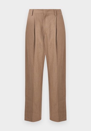 Junger Mann steht mit den Händen in den Taschen, trägt ein braunes, schwarz-weiß gestreiftes kurzärmeliges Hemd, weite beigetöne Hosen und dunkle Sandalen.