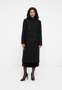 Cappotto lungo in misto lana nera doppiopetto con grandi bottoni frontali e colletto classico, abbinato a una gonna nera plissettata e tacchi neri.