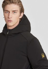 Zwarte capuchonjack met een gladde textuur, voorzien van een voorrits en een logo-opdruk op de linkermouw. Eenvoudig ontwerp en een getailleerde pasvorm.