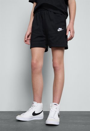 Person trägt schwarze Nike-Shorts und weiße Nike High-Top-Sneaker und steht auf grauem Boden vor einer hellen Wand.